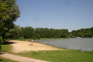 Sandstrand am See mit Badegästen, Baumreihe am Ufer und ruhigem Wasser im Hintergrund.