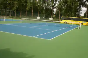Tennisplatz auf dem Campinggelände mit Netz und Linienmarkierungen, im Hintergrund ein gelber Bus und Bäume.