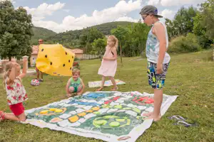 Familie spielt auf einer Campingwiese mit großem Spielteppich, im Hintergrund Bäume und Hügel sowie ein Wohnwagen.