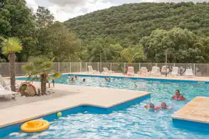 Schwimmbad auf dem Campingplatz mit mehreren Liegen, Sonnenschirmen und Palmen am Beckenrand, im Hintergrund bewaldete Hügel.