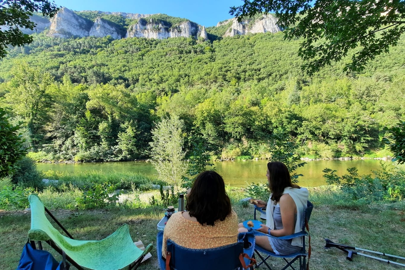 Campingplatz am See mit zwei Personen auf Klappstühlen, Blick auf grünen Wald und Felswände in den Bergen.