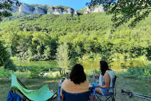 Campingplatz am See mit zwei Personen auf Klappstühlen, Blick auf grünen Wald und Felswände in den Bergen.