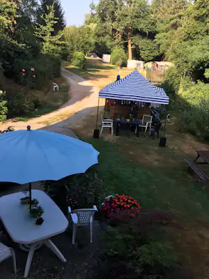 Terrasse mit blauem Sonnenschirm, Tisch und Stühlen am Campingplatz, im Hintergrund Zelt und Wege zwischen Bäumen.