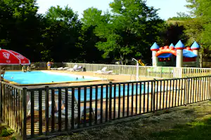 Schwimmbad mit blauem Wasser, Sonnenschirmen, Liegestühlen und einem Hüpfburg-Spielbereich hinter dem Zaun.