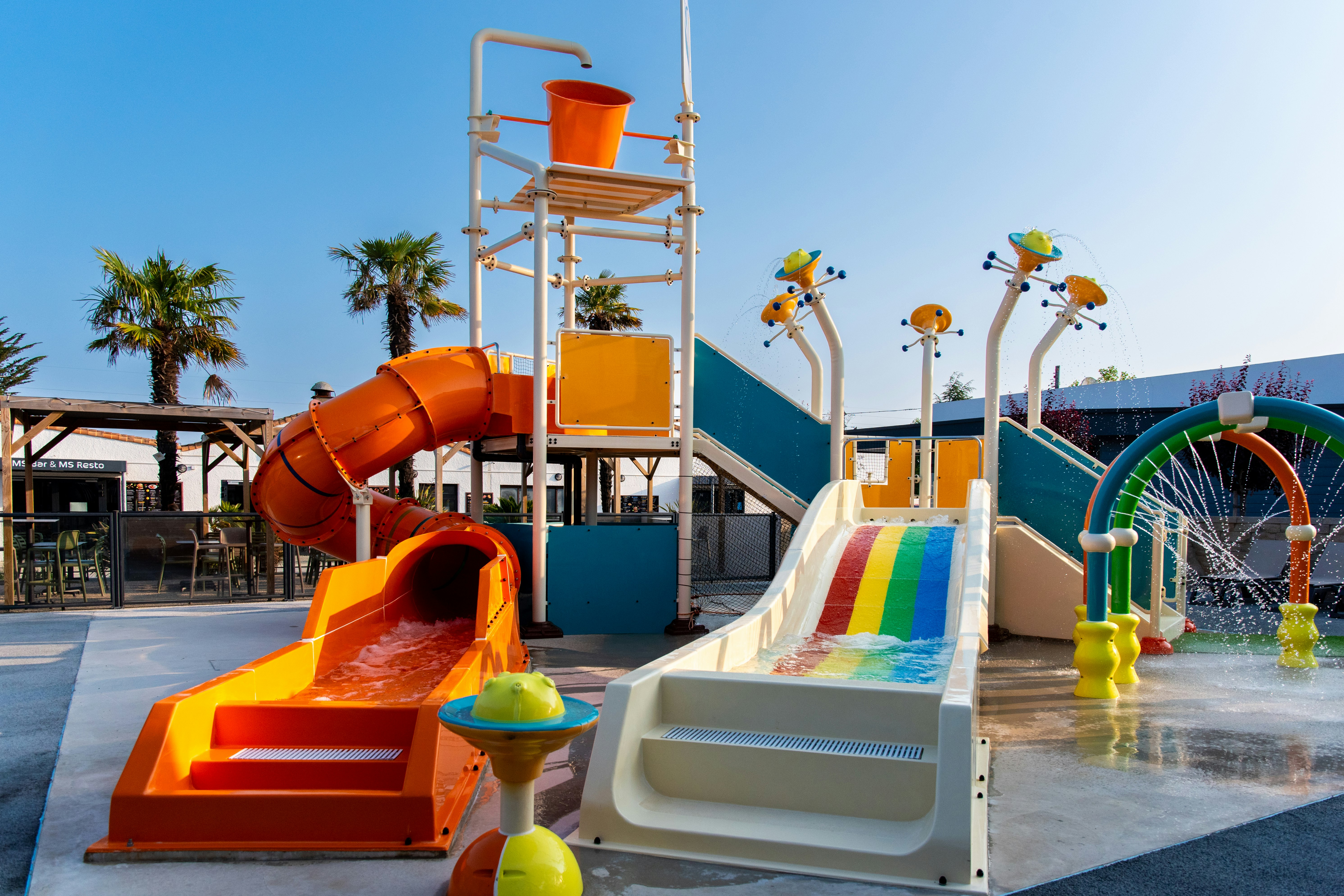 Wasserspielbereich mit bunten Rutschen und Spielzeug, daneben ein orangefarbenes Wasserbecken und Palmen vor blauem Himmel.