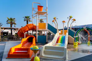 Wasserspielbereich mit bunten Rutschen und Spielzeug, daneben ein orangefarbenes Wasserbecken und Palmen vor blauem Himmel.