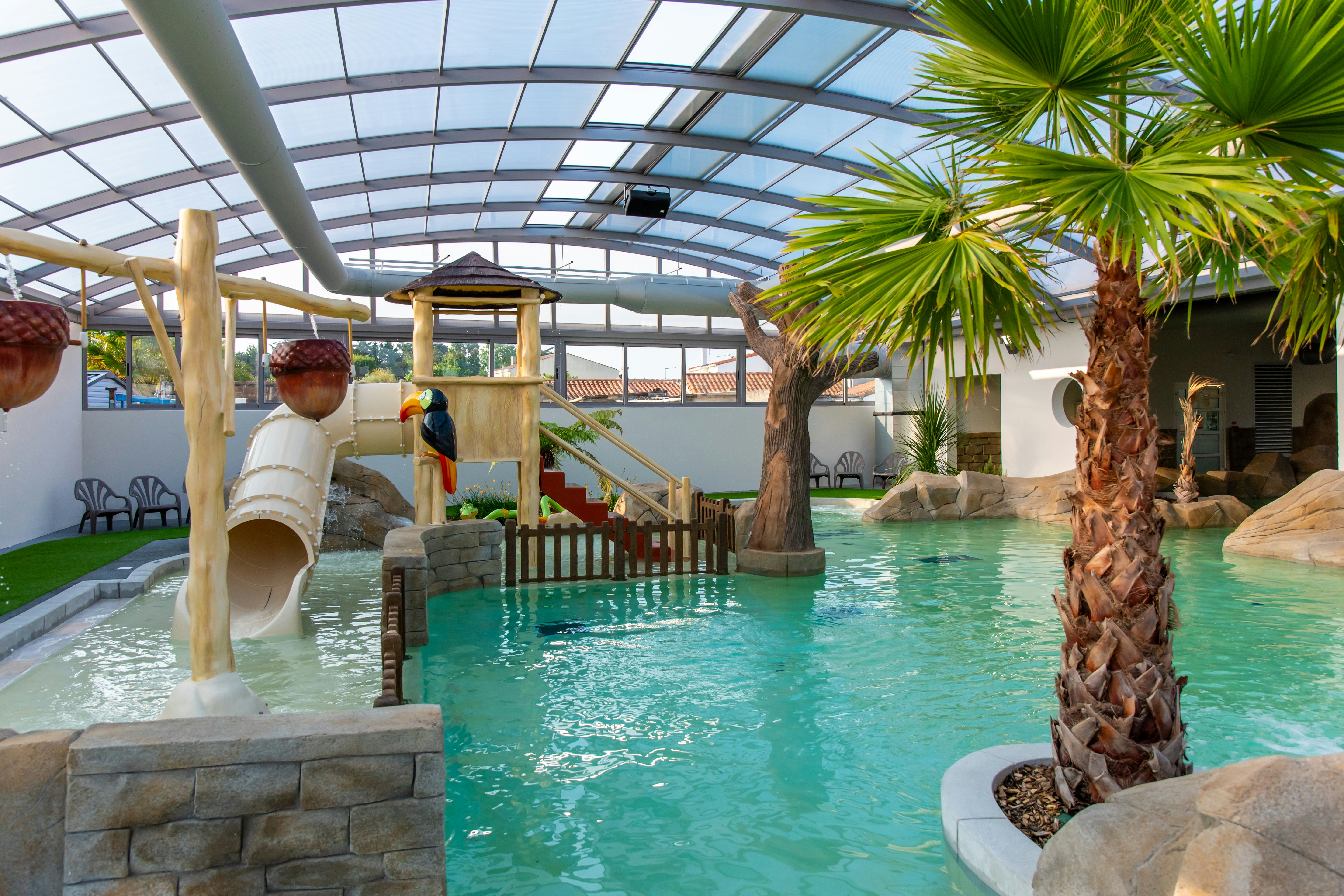 Innenpool mit Glasdach, Palmen und Steinmauer; Wasserrutsche und Spielbereich mit Holzhaus im Hintergrund.