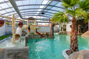 Innenpool mit Glasdach, Palmen und Steinmauer; Wasserrutsche und Spielbereich mit Holzhaus im Hintergrund.