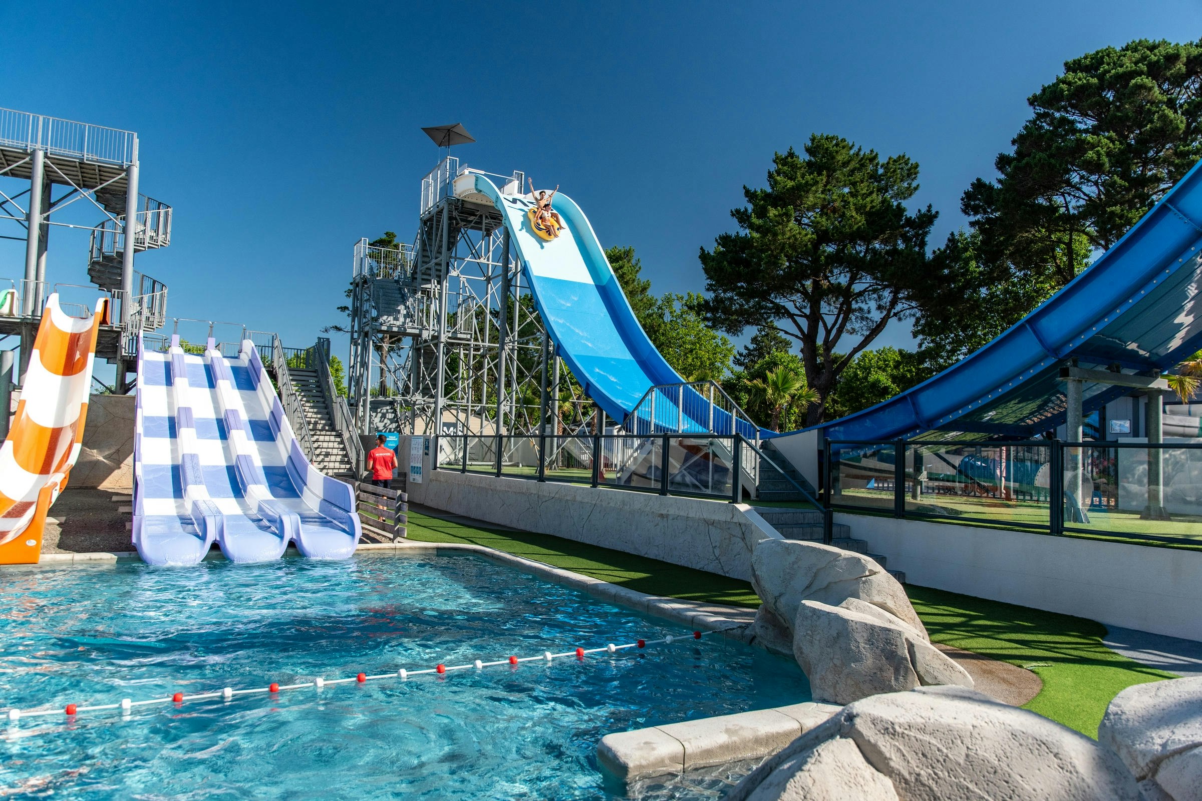 Blauer Wasserrutschenbereich mit Planschbecken, Sicherheitsgeländer und Liegen am Campingplatz, im Hintergrund Bäume und Gebäude.
