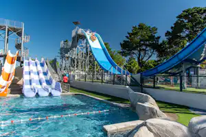 Blauer Wasserrutschenbereich mit Planschbecken, Sicherheitsgeländer und Liegen am Campingplatz, im Hintergrund Bäume und Gebäude.