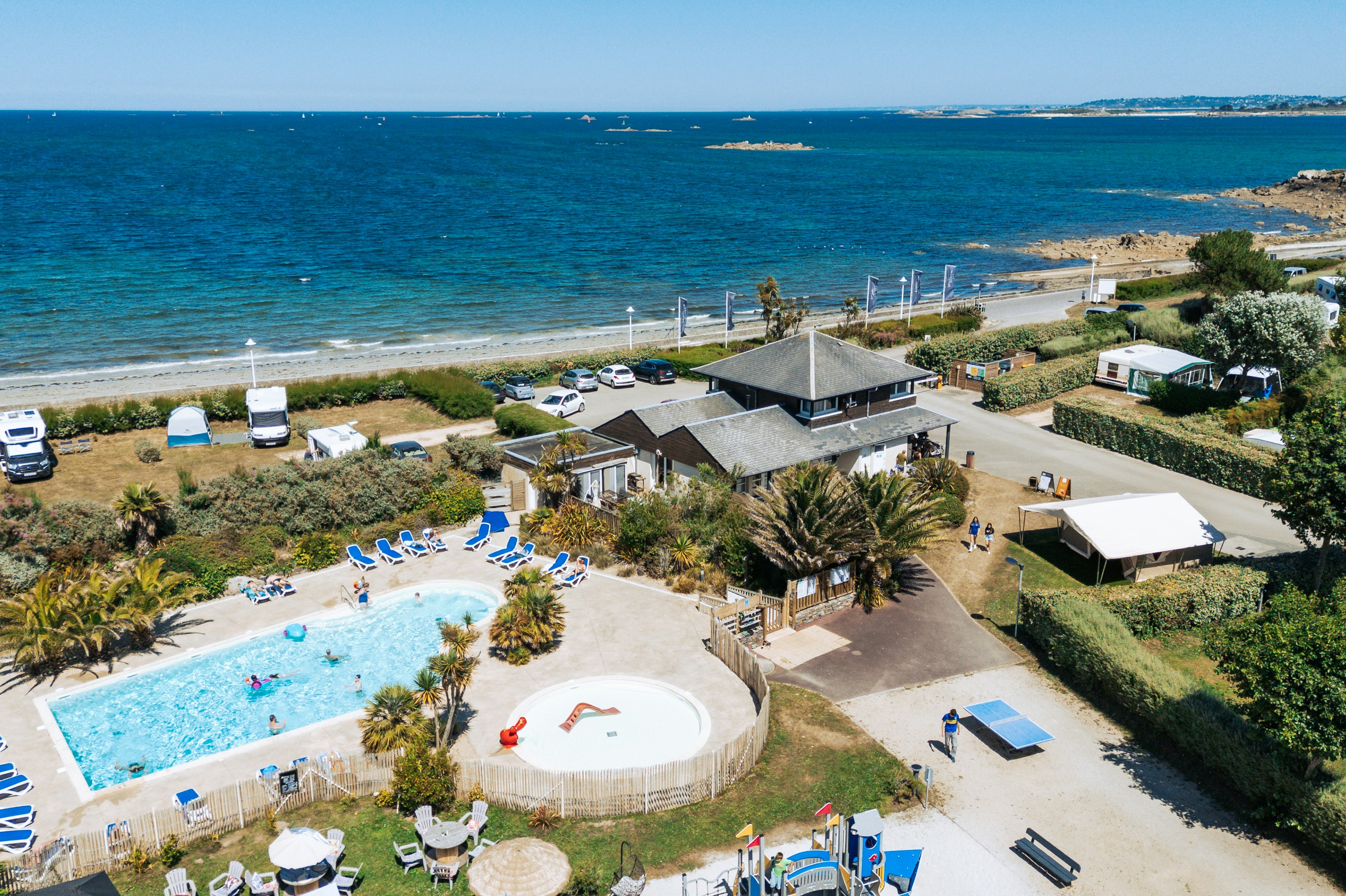 Campinganlage am Meer mit Pool, Sonnenschirmen, Stellplätzen und Wohngebäuden am Strand, im Hintergrund Schiffe auf dem Wasser.