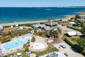 Campinganlage am Meer mit Pool, Sonnenschirmen, Stellplätzen und Wohngebäuden am Strand, im Hintergrund Schiffe auf dem Wasser.