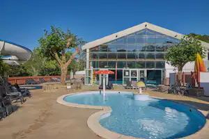 Schwimmbad mit rundem Becken, Sonnenschirmen und Liegen vor einem modernen Gebäude mit Glasfront und Bäumen.