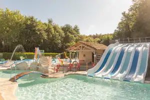 Kinderpool mit blauer Rutsche, Holzdeck und Liegen am Rand, im Hintergrund Spielbereich und Bäume im Grünen.