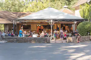 Restaurant-Terrasse auf dem Campingplatz mit Gästen an Tischen, Steinsockel und Sonnenschirm vor Gebäuden und Bäumen.