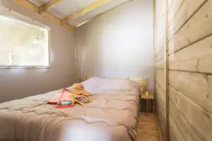 Bett in einem Holzschlafzimmer im Campinghaus, mit Nachttisch, Lampe und Fenster im Seitenbereich.