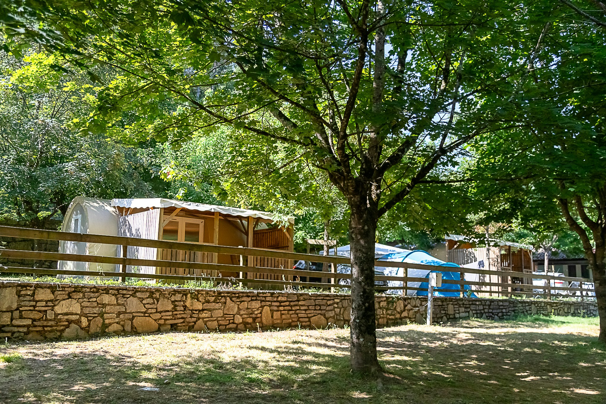 Wohnwagen auf einem Campingplatz zwischen Bäumen, mit Holzgeländer, Steinmauer und Grasfläche im Vordergrund.