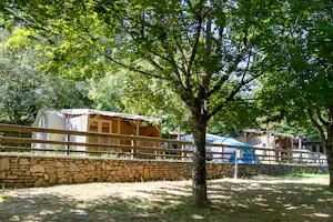 Wohnwagen auf einem Campingplatz zwischen Bäumen, mit Holzgeländer, Steinmauer und Grasfläche im Vordergrund.