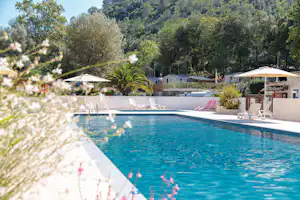 Schwimmbecken mit Liegen am Campingplatz, im Hintergrund Bäume und Gebäude, Sonnenschirme am Beckenrand.