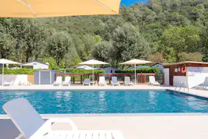 Schwimmbad mit Liegestühlen und Sonnenschirmen, im Hintergrund Bungalows und dicht bewachsene Hügel.