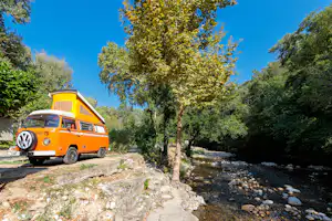 Oranger VW-Camper steht auf einem Kiesweg am Bach, umgeben von Bäumen und Felsen unter blauem Himmel.