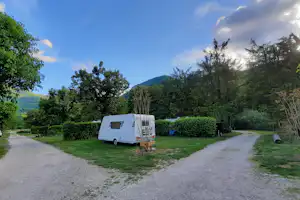 Weißes Wohnmobil auf einem Stellplatz im Grünen, Kiesweg, Hecken und Bäume vor grünen Hügeln und blauem Himmel.