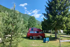 Ein rotes Wohnmobil auf einem Stellplatz im Grünen, mit Tisch und Stühlen, Bäumen und Blick auf die Berge.