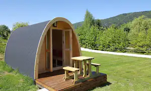 Holz-Kuppelzelt mit Eingang und Fenstern, davor eine Holzterrasse mit Tisch und Bänken, umgeben von Grün und Bäumen.