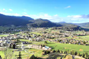 Blick auf ein Dorf im Tal mit grünen Wiesen, Häusern und Bergen im Hintergrund.