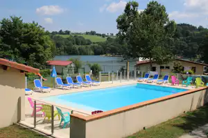 Blick auf den Camping-Pool mit Liegestühlen und Sonnenschirmen, dahinter ein See und grüne Hügel.