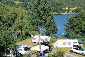 Campingplatz am See mit Wohnwagen, Zelten und Auto auf dem Stellplatz, umgeben von Bäumen und grünen Wiesen.