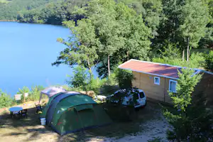 Grünes Campingzelt am Seeufer, daneben ein Wohnwagen und ein kleines Holzhaus im Grünen.