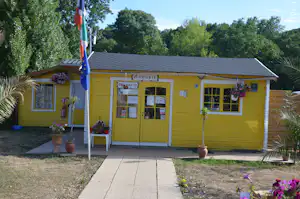 Gelbes Campinghaus mit Eingangstür und Fenstern, davor Gehweg und Blumenbeete, im Hintergrund Bäume und Flagge.