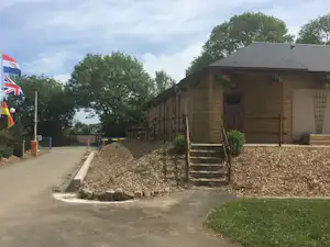 Holzhütte auf einem Campingplatz mit Treppe, Sitzbank und Heckenbeet neben dem Weg, im Hintergrund Bäume und Flaggen.
