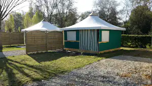 Campingzelt mit gestreifter Markise auf einem Stellplatz, umgeben von Bäumen und einem Holzsichtschutz.
