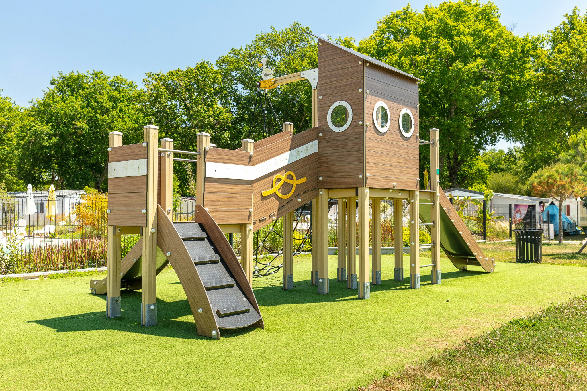 Spielplatz mit hölzernem Kletterturm und Rutsche auf dem Campingplatz, umgeben von Rasen und Bäumen.
