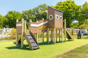 Spielplatz mit hölzernem Kletterturm und Rutsche auf dem Campingplatz, umgeben von Rasen und Bäumen.