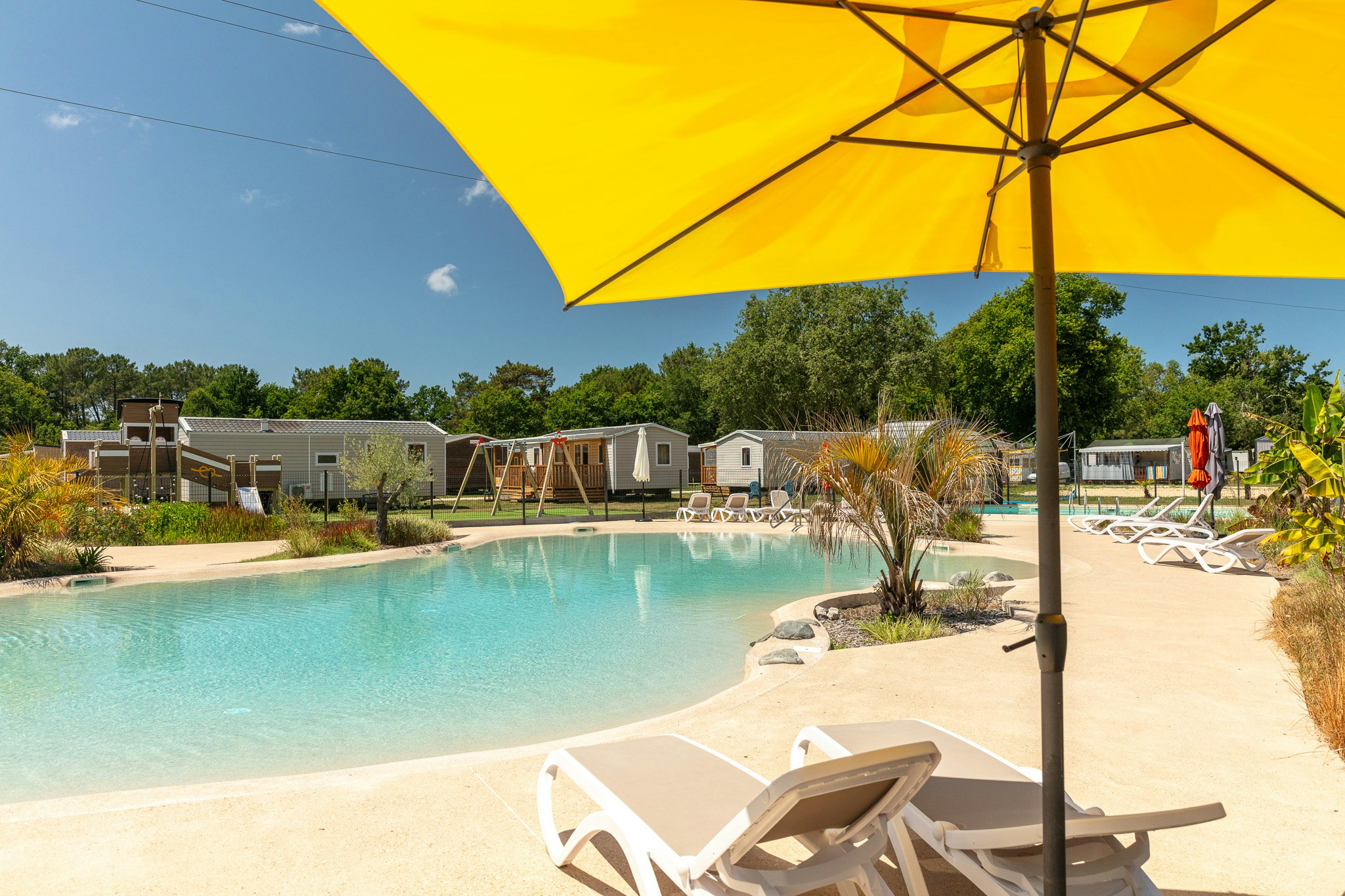 Pool am Campingplatz mit Sonnenschirm, Liegen und gepflegtem Garten; im Hintergrund Wohnwagen und kleine Gebäude.