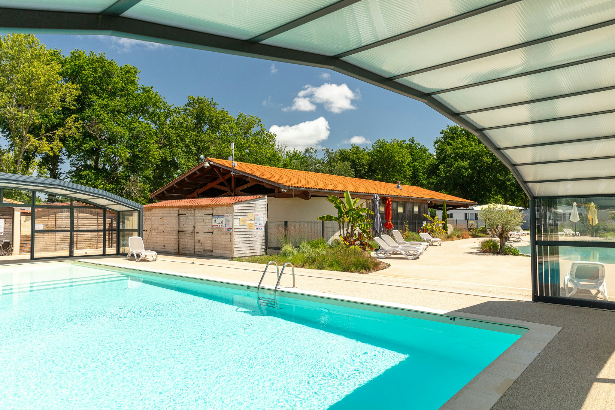 Beheizter Camping-Pool unter einem Glasdach, daneben Liegen und Sonnenschirme, im Hintergrund Gebäude und Bäume.