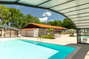 Beheizter Camping-Pool unter einem Glasdach, daneben Liegen und Sonnenschirme, im Hintergrund Gebäude und Bäume.