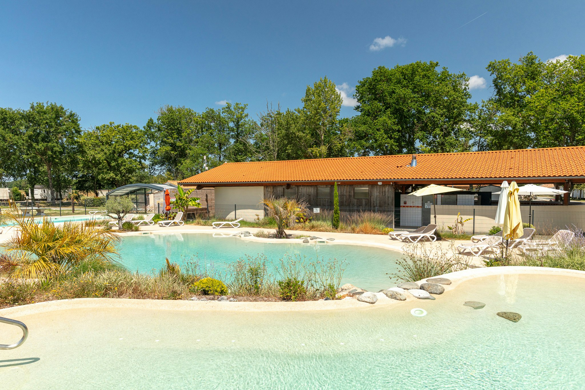 Türkisfarbenes Camping-Poolbecken mit Liegen und Sonnenschirmen vor einem Gebäude mit orangefarbenem Dach und Bäumen.