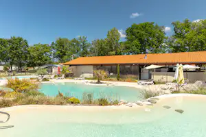 Türkisfarbenes Camping-Poolbecken mit Liegen und Sonnenschirmen vor einem Gebäude mit orangefarbenem Dach und Bäumen.