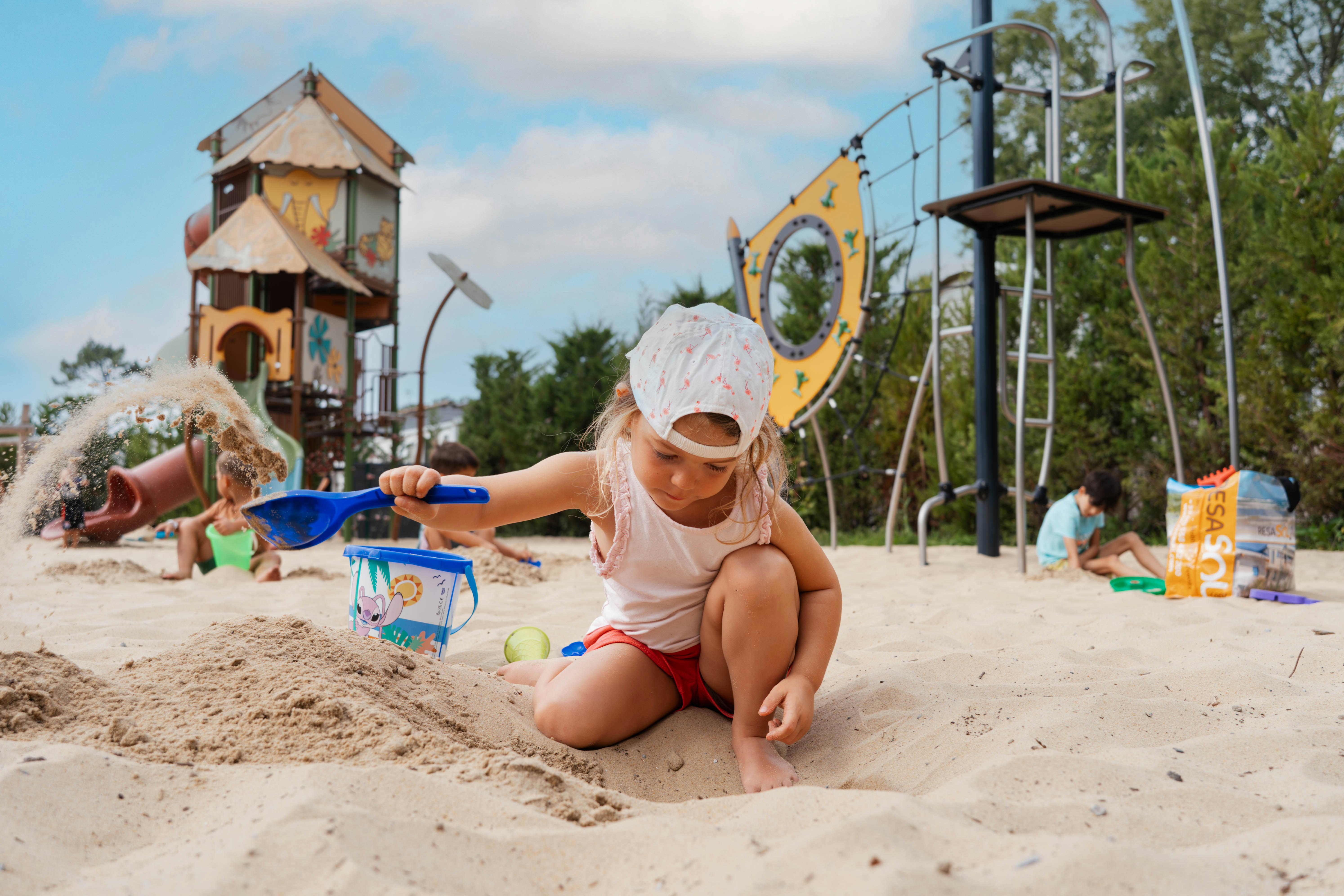 Kleines Kind im Sandkasten mit Schaufel, Spielzeug und Wasserspiel am Campingplatz, im Hintergrund Spielhaus und Klettergerüst.