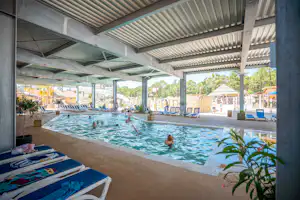 Überdachtes Schwimmbad mit blauem Wasser, mehreren Liegen und Sonnenschirmen unter einem Metall-Dach.