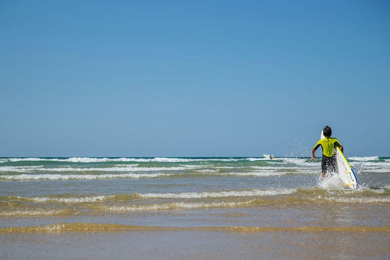 Ein Surfer mit Surfbrett steht im flachen Wasser am Strand, Wellen rollen an der Küste entlang.