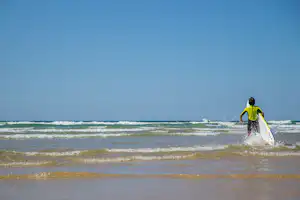 Ein Surfer mit Surfbrett steht im flachen Wasser am Strand, Wellen rollen an der Küste entlang.
