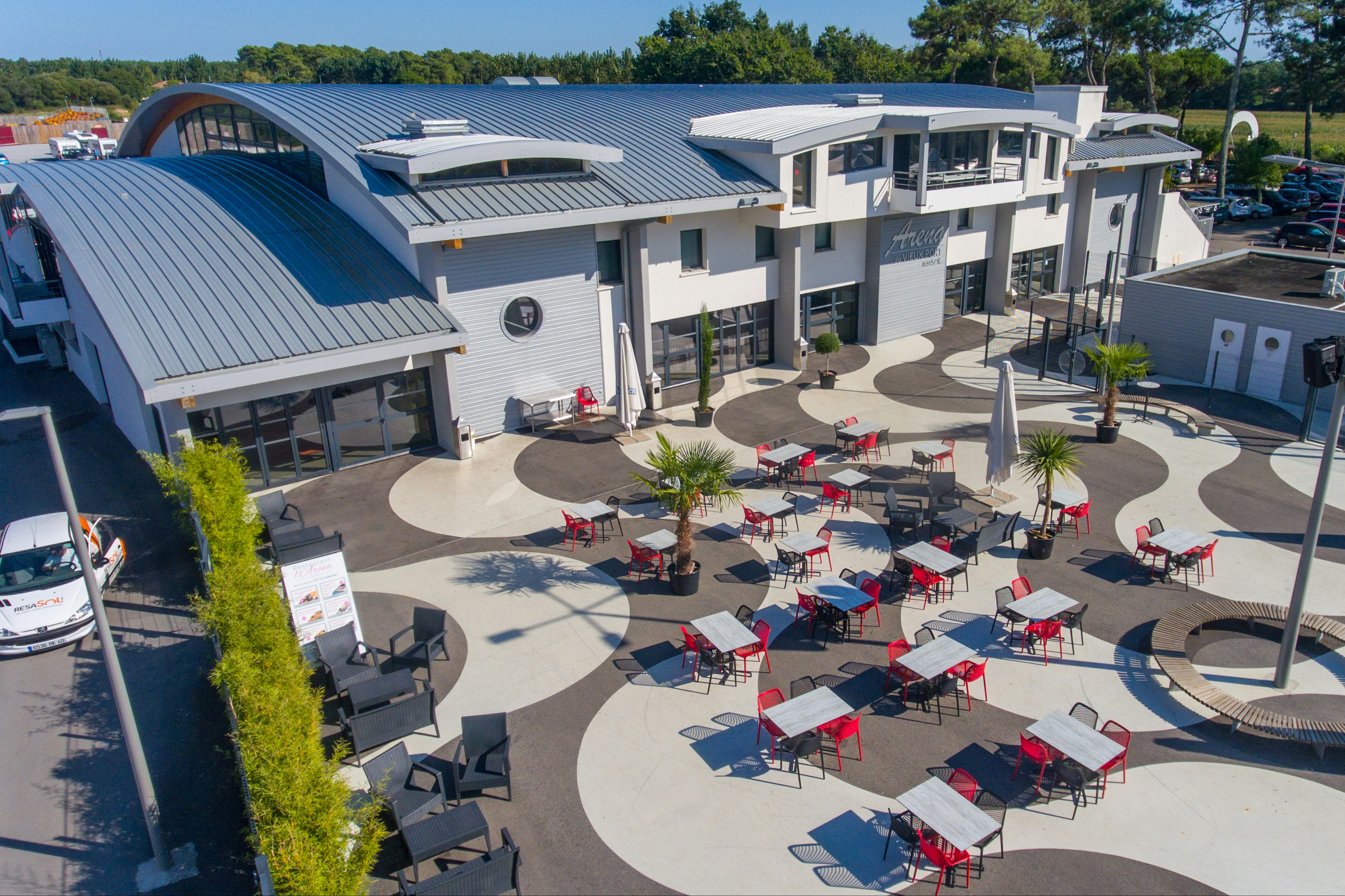 Innenhof eines Campingplatzes mit Reihen von Stellplätzen, Sonnenschirmen und Tischen vor modernen Gebäuden und Bäumen.