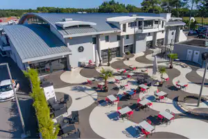 Innenhof eines Campingplatzes mit Reihen von Stellplätzen, Sonnenschirmen und Tischen vor modernen Gebäuden und Bäumen.