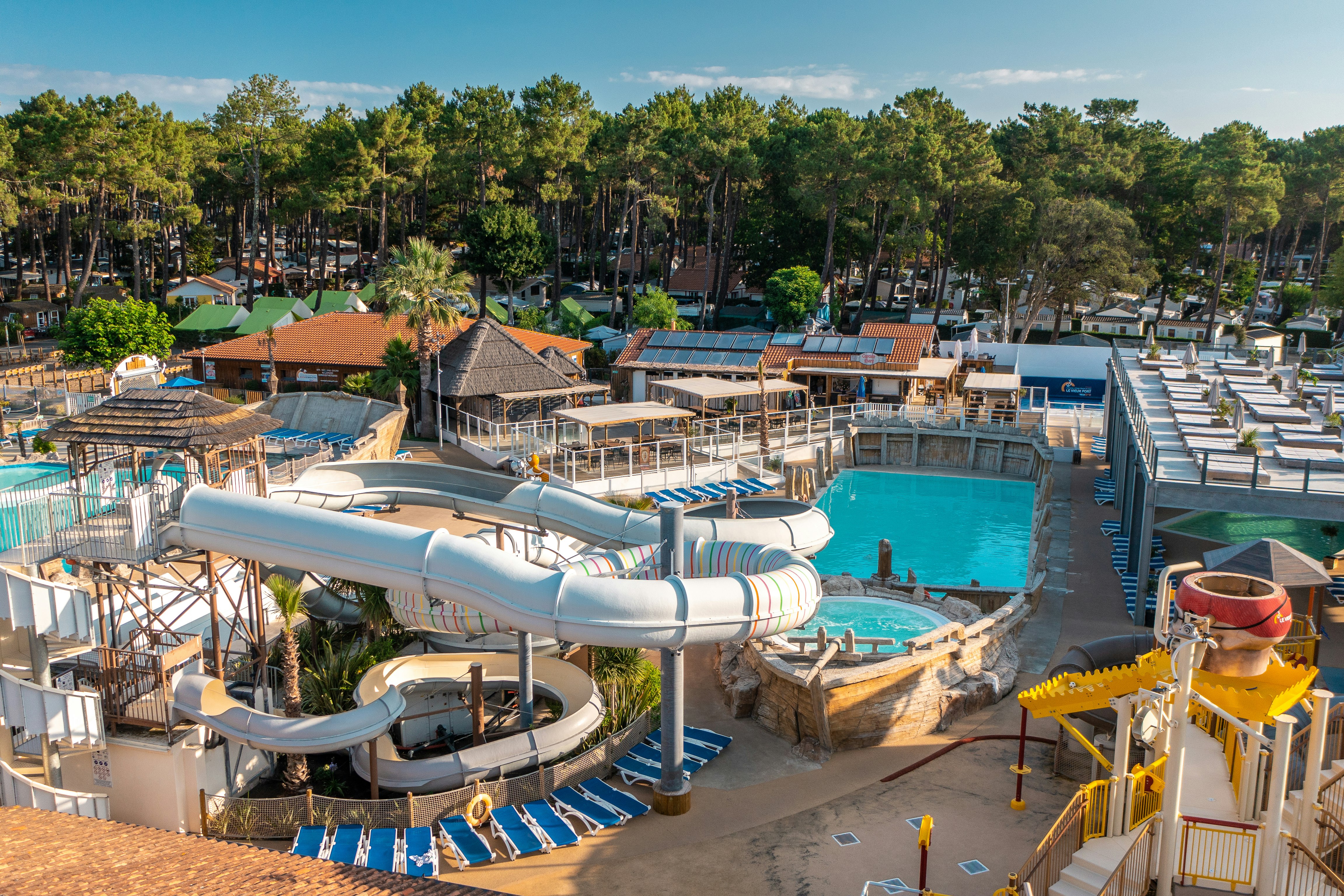 Großer Campingplatz mit Poolbereich, Liegestühlen, Sonnenschirmen und mehreren Wasserrutschen, umgeben von Bäumen und Wohnwagen.