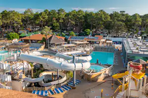 Großer Campingplatz mit Poolbereich, Liegestühlen, Sonnenschirmen und mehreren Wasserrutschen, umgeben von Bäumen und Wohnwagen.